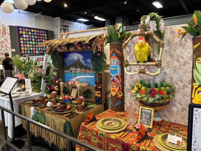 Two tropical-themed table setting displays, one featuring a view of what looks to be Fiji, and both with tiki totems. On the right is a stuffed parrot modeled on the ones from Disneyland's Tiki Room.