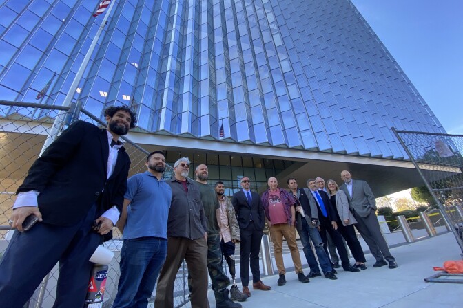 A group of 12 people stand in a semicircle front of a tall glass building with an American flag out front