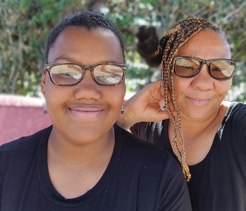 Two women pose for a photograph. Both are smiling, wearing black t-shirts and glasses. On the right, the adult woman curls her knuckles against her jaw in a relaxed pose.