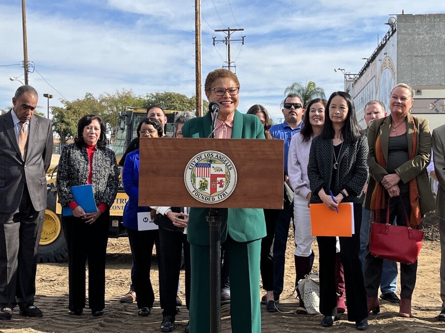 L.A. Mayor Karen Bass, a woman with light brown skin and a close-cropped haircut, stands in front of a wooden podium with the logo of the Los Angeles Mayor's office on it at a dirt-covered outdoor construction site. Eleven people, both men and women of various ages and ethnicities, stand behind her.