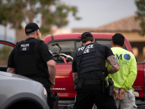 Two police/ICE officers in front of a red truck arresting something in a bright yellow shirt