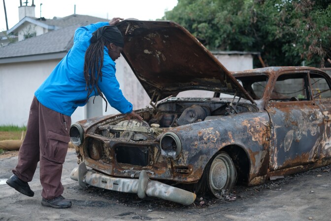 A man with dark skin tone, long black dreadlocks and a blue jacket looks under the hood of a burned car. 