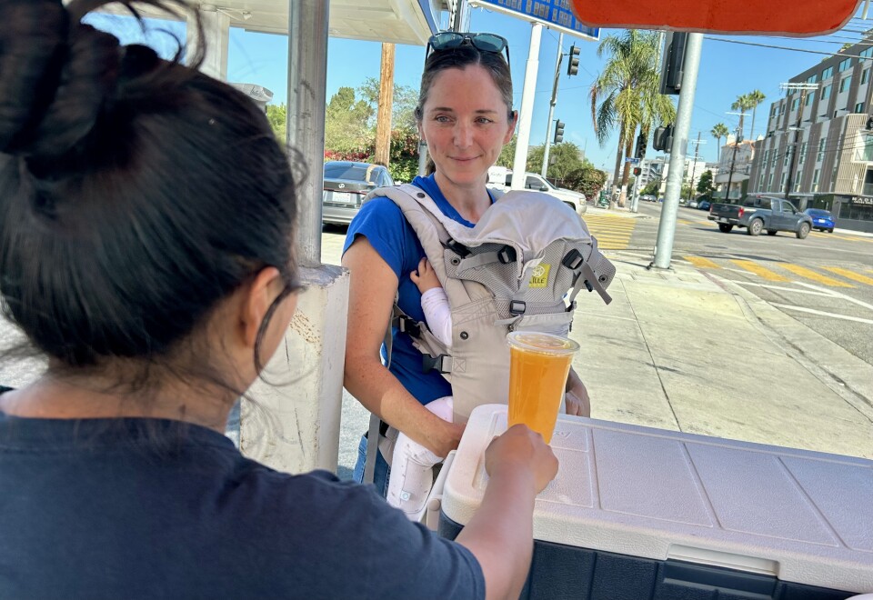 A light skinned woman holding a baby in a carrier is talking to another woman with dark hair whose back is to the camera. There is a plastic cup og orange juice on an ice chest in front of them.