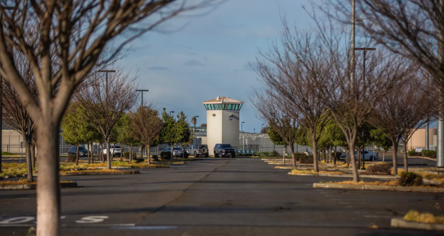 A watchtower in the background, with cars on the other side of a chain-link fence. In the foreground, the watchtower is framed by trees.