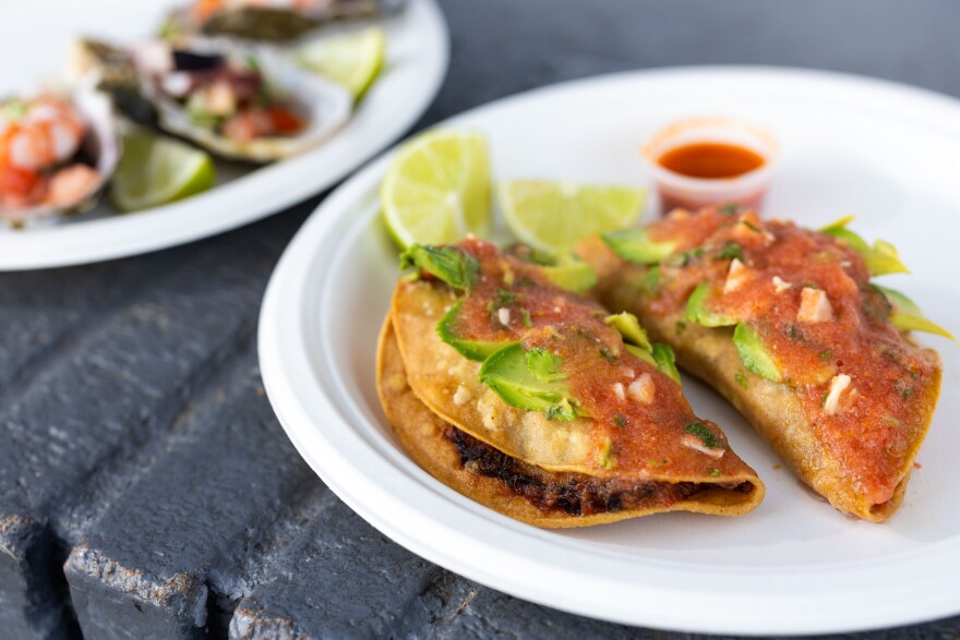A close-up of two folded fried tacos reveals their cooked filling peeking out slightly. Both tacos are topped with a red sauce and garnished with a slice of green avocado. They are served on a white paper plate, accompanied by two lime wedges and a small plastic container filled with red salsa.