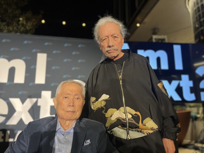 A Japanese American man and Latino man pose together in front signs that read "Am I Next?"