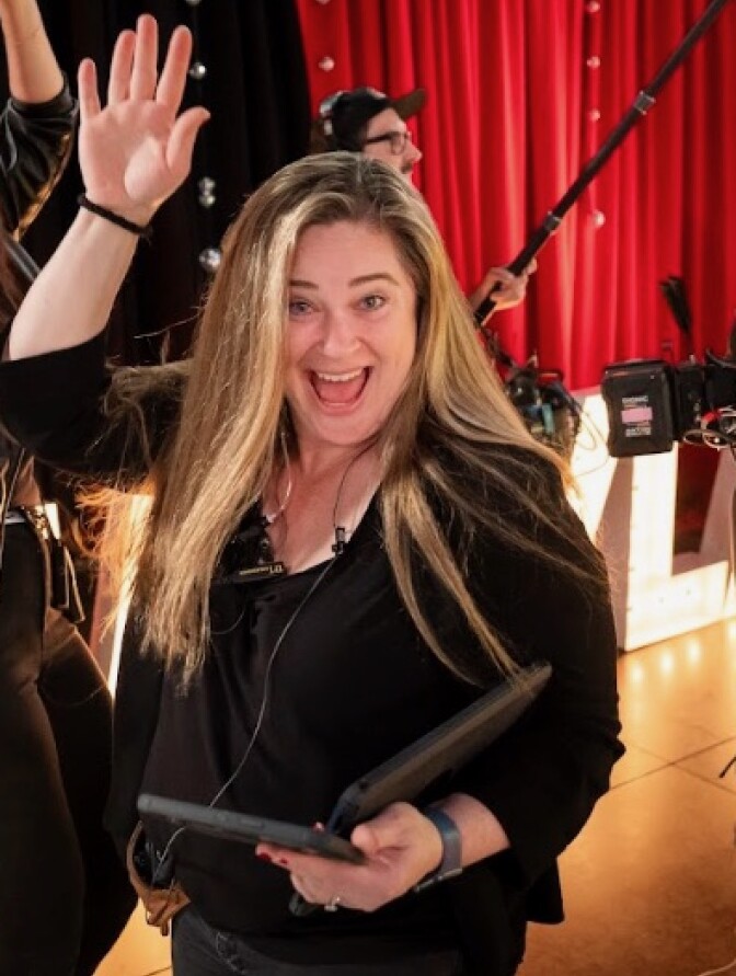 A white woman with long blonde hair mugs for the camera while on a the set of a TV show. 