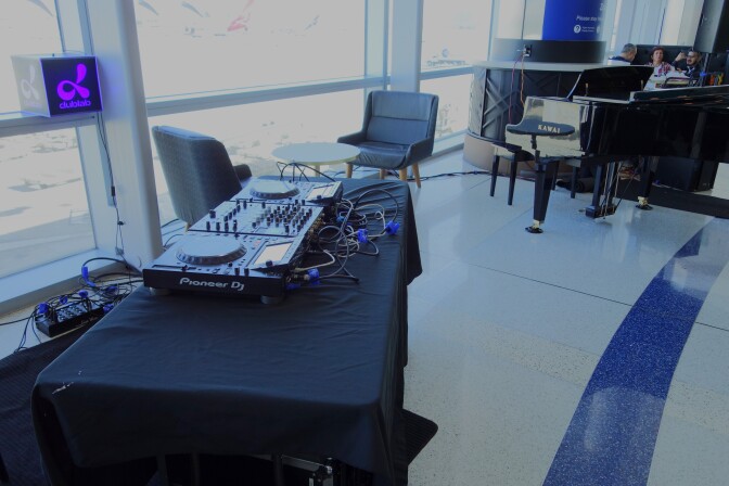 An empty makeshift DJ booth with Pioneer DJ decks. A piano, empty chairs and several large windows are in the background.