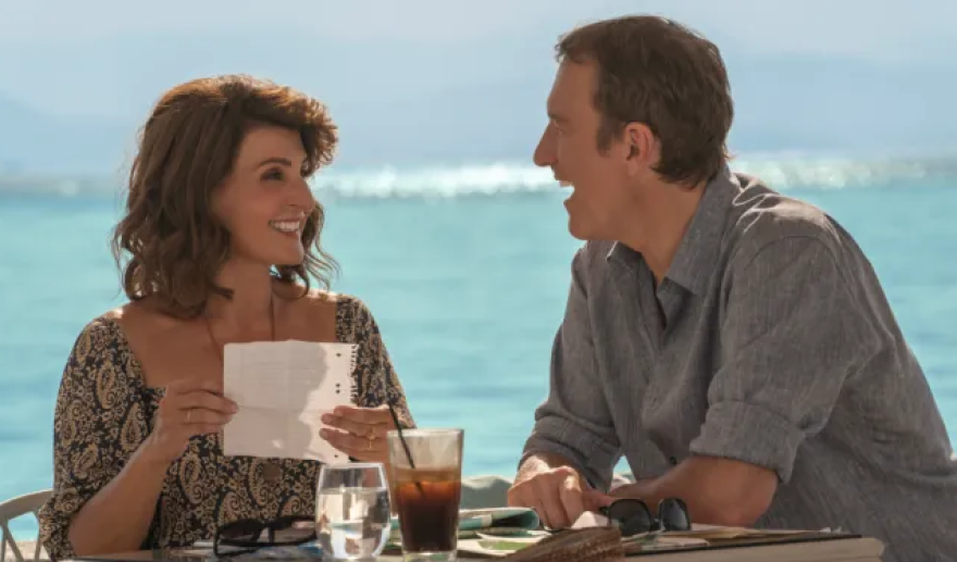 Nia Vardalos and John Corbett sit a table oceanside, bright blue waves crashing  behind them. Vardalos looks at Corbett smiling. She is holding a piece of paper that seems to be a page ripped from a notebook. Corbett looks at her, his mouth open as if laughing. 