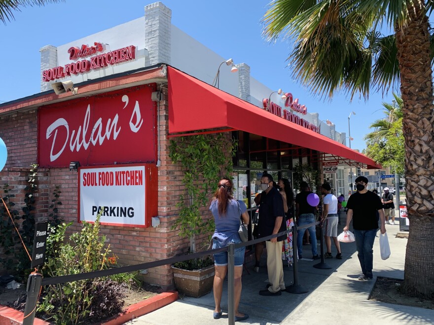 People wait outside a brick building with a red and white awning and a red sign with the restaurant's name