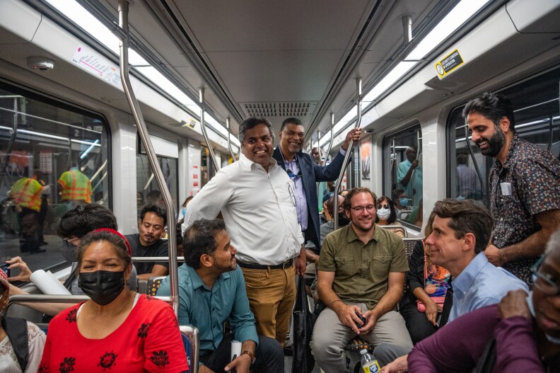 A Metro train car is filled with a diverse group of riders. Every seat is taken and several people are standing. Nearly everyone is smiling. A few still wear medical masks, a sign of the lingering COVID-19 pandemic.