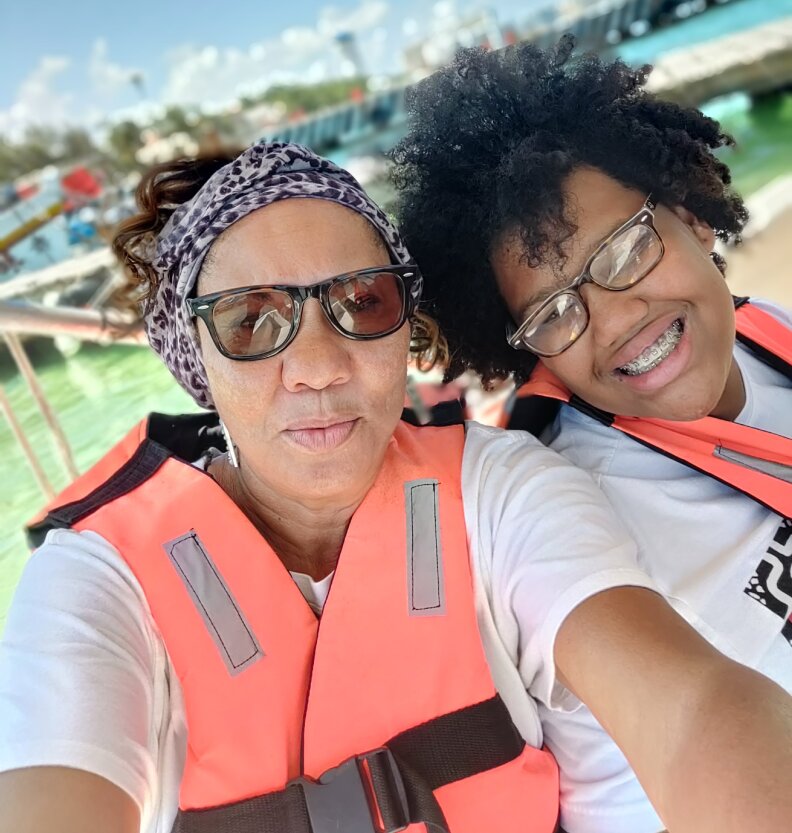 Two women pose for a photograph near what looks like a marina or harbor. Both are wearing salmon-colored life jackets and glasses. The younger girl in the photograph smiles to reveal braces on her teeth.