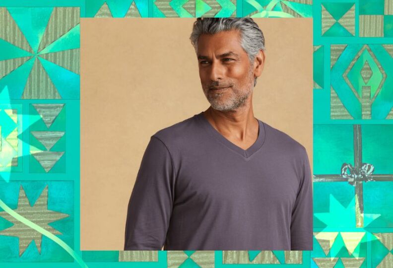 A photograph of a man wearing a classic, long sleeved V-neck T-shirt in a gun metal gray. The image is set against a decorative green background accented with stars, gift boxes and strands of ribbon.