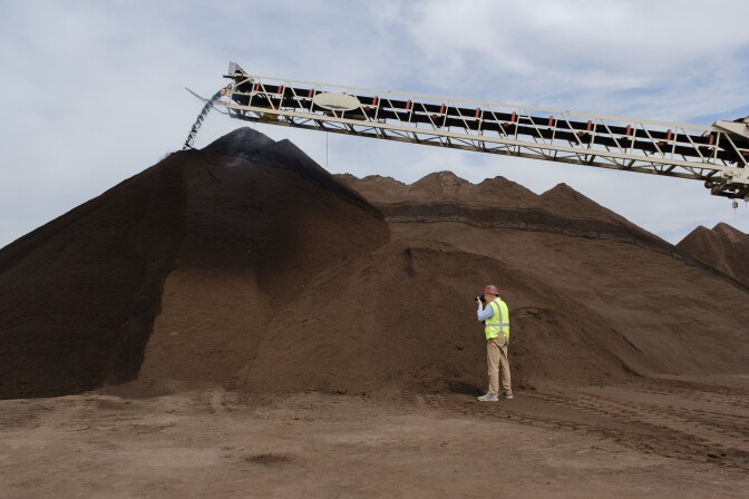 A man in a neon safety vest takes an image of a large pile of rich brown compost that towers more than a dozen feet over his head.