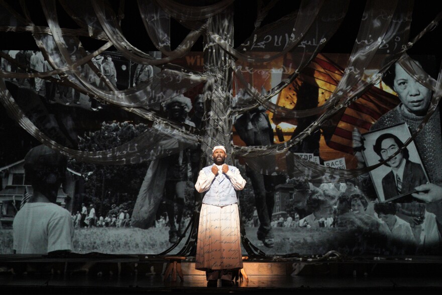 A photo of tenor Jamez McCorkle as Omar in LA Opera's 2022 West Coast premiere of "Omar." He is dressed in a white costume that has Arabic script written on it. He stands before a collage of images from the American Civil Rights movement.