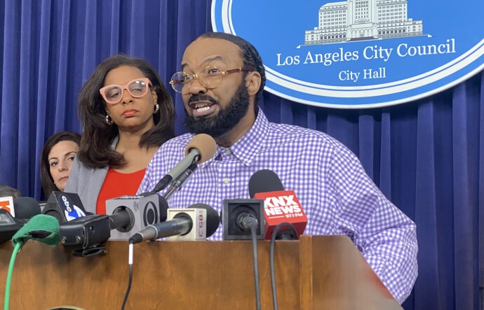A Black man speaks at a podium inside Los Angeles City Hall.