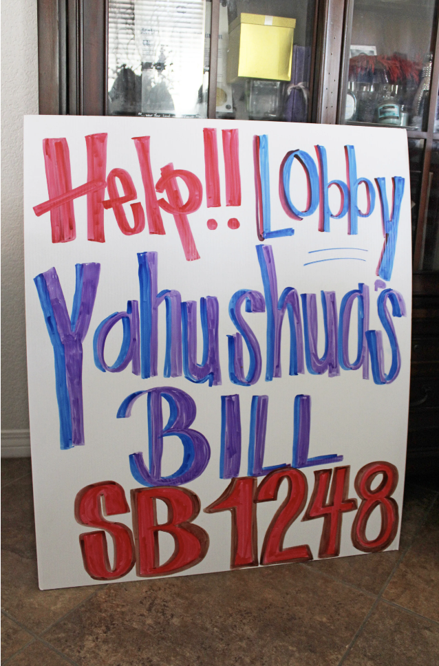 A sign sits in a living room and reads, in blue and white hand-painted letters, "Help!! Lobby Yahushua's Bill SB 1248"
