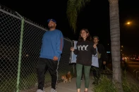 Three people with medium skin tone and a small dog on a leash walk down a sidewalk past a metal fence and palm tree at night.