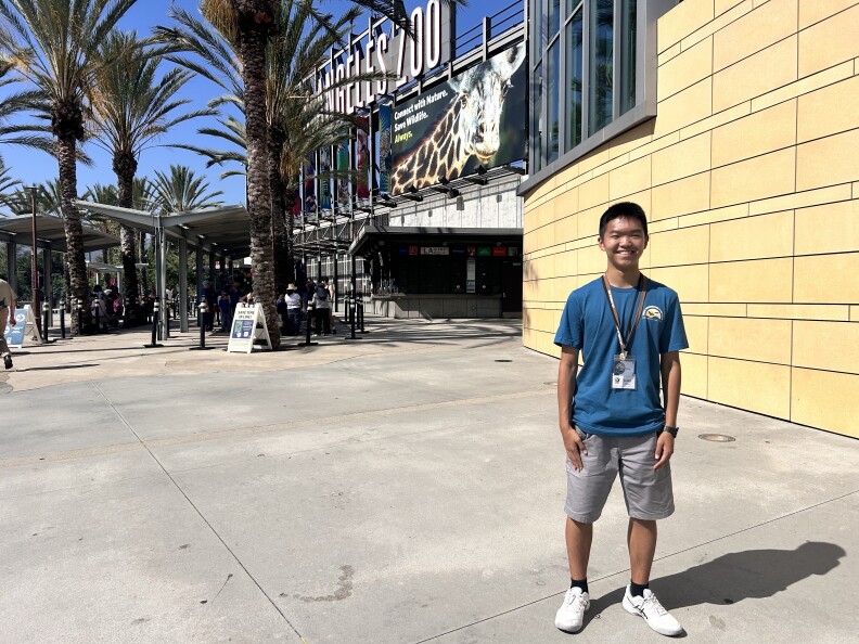A teenage boy in a blue t-shirt and gray shorts is smiling in front of the exterior of the Los Angeles Zoo. Both hands are to his side and he's wearing a lanyard with a pair of name tags pinned to it.