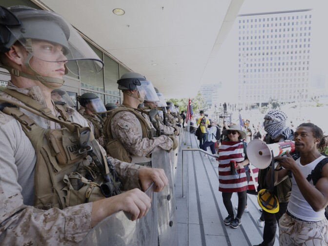 A row of troops hold shields outside a building as people gather on the steps. 