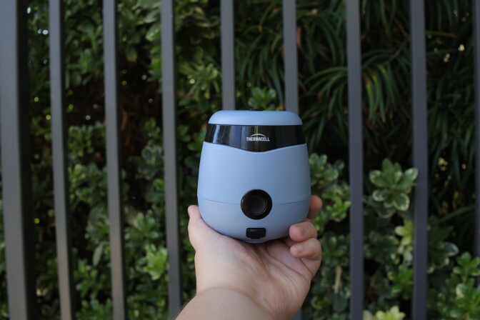 A hand with a light skin tone holds up a round light blue device in front of green plants and black bars. The device has a black button near the button with the company name Thermacell above it.