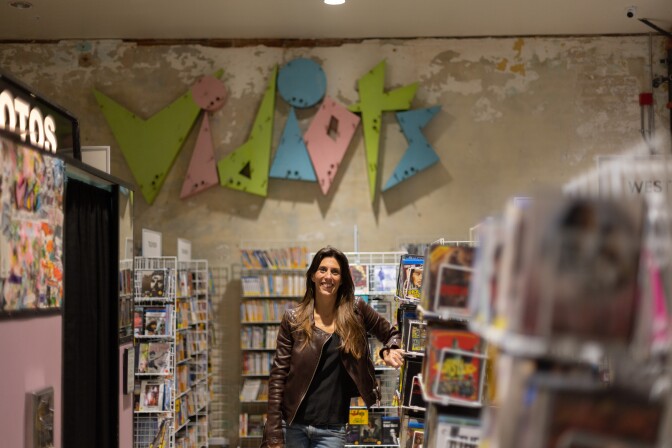 A feminine presenting person with light skin tone and light brown hair wearing a brown leather jacket and jeans stands leaning on a stand full of magazines. Behind them on the wall is a colorful sign that reads "Vidiots."