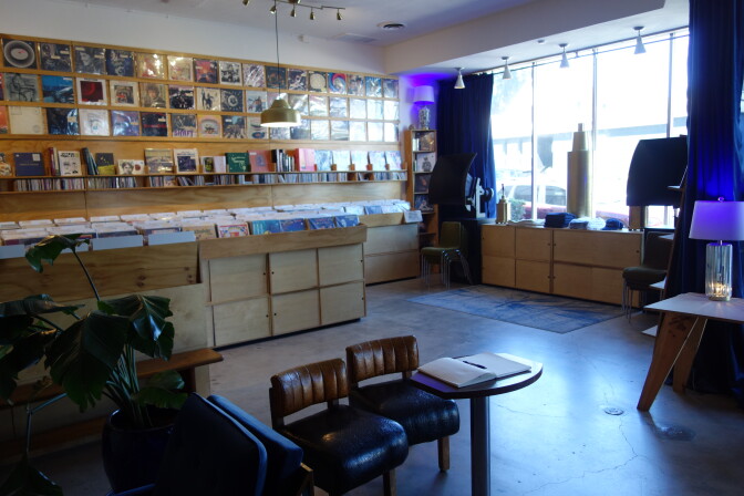 Interior of a record shop with vinyl albums displayed on the back wall and in wooden bins, a small round table with two leather chairs in the foreground, and large windows with blue curtains along the right side.