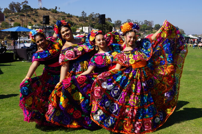 A group of four medium-skinned woman in brightly colored dance dresses with flowers in their hair. 