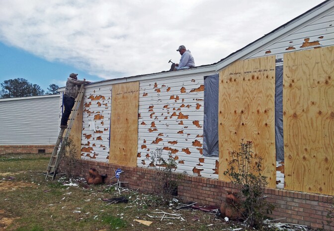 Two men, one on a ladder and the other on a roof. Three large wood panels board up portions of a the home that has been damaged.