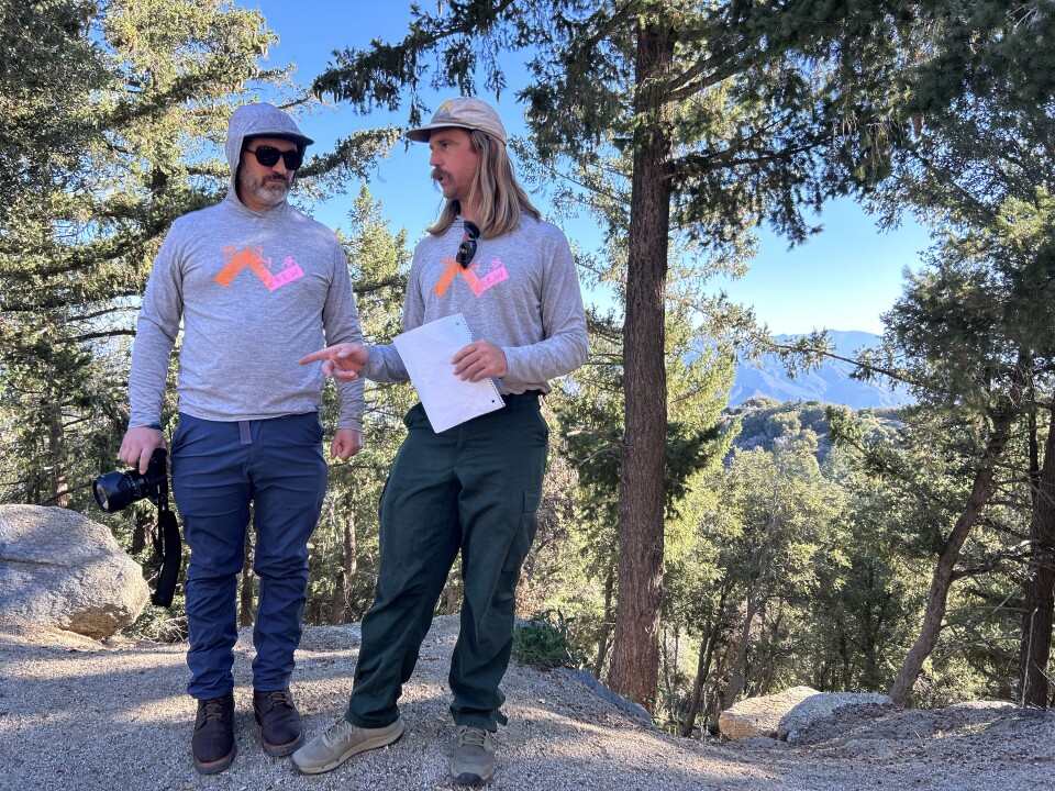 Two middle-aged white men wearing the same gray sweatshirt and work pants and boots talk with sun-glowing pines in the background. 