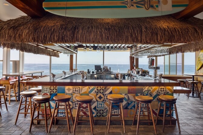 A bar overlooking the Pacific ocean: There are wooden stools lining the bar and high top tables, all positioned for a view of the waves. The decor is a little bit tiki (there's a grass fringe skirt rings the cabinetry above the bar), but also a little beachy (there's are surfboards on the wall and above the bar) and colorful tiles accent the front of the bar.