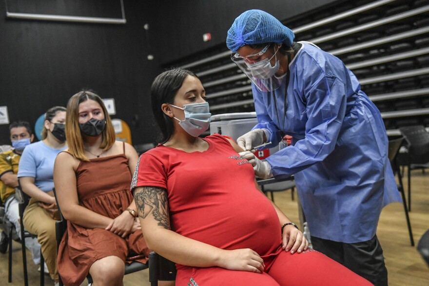 A pregnant woman on a red dress wears a surgical mask as a health care worker in protective gear gives her a shot her upper left arm.