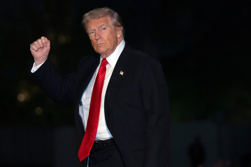 President Trump, a man with light skin tone wearing a black suit with a red tie, holds his right fist up as he looks towards the camera.
