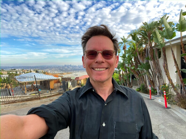A man with a light skin tone smiles and takes selfie while standing outside. In the background is the neighborhood landscape with green plants, a bright blue and cloudy sky, and a view of Los Angeles towers in the distance.