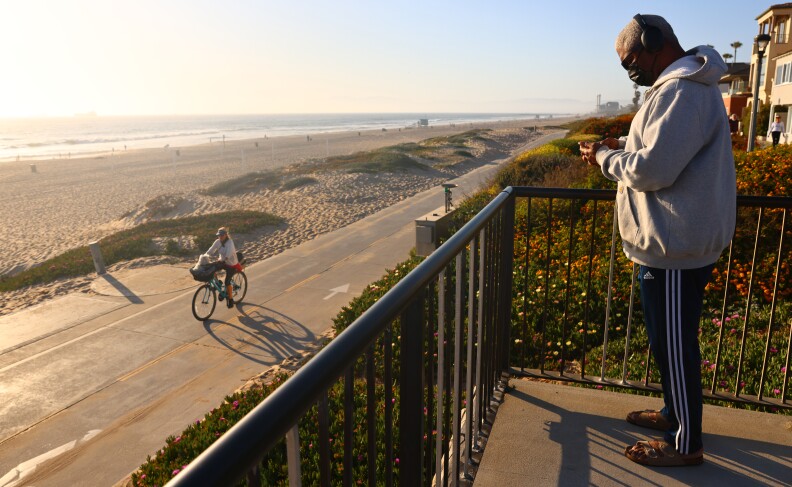 A man who appears to be Black and in middle age checks his phone while standing on a balcony overlooking a bicycle path along a beach. The sun appears the be setting. The beach is a very long, continuous stretch of sand. The ocean can be seen.