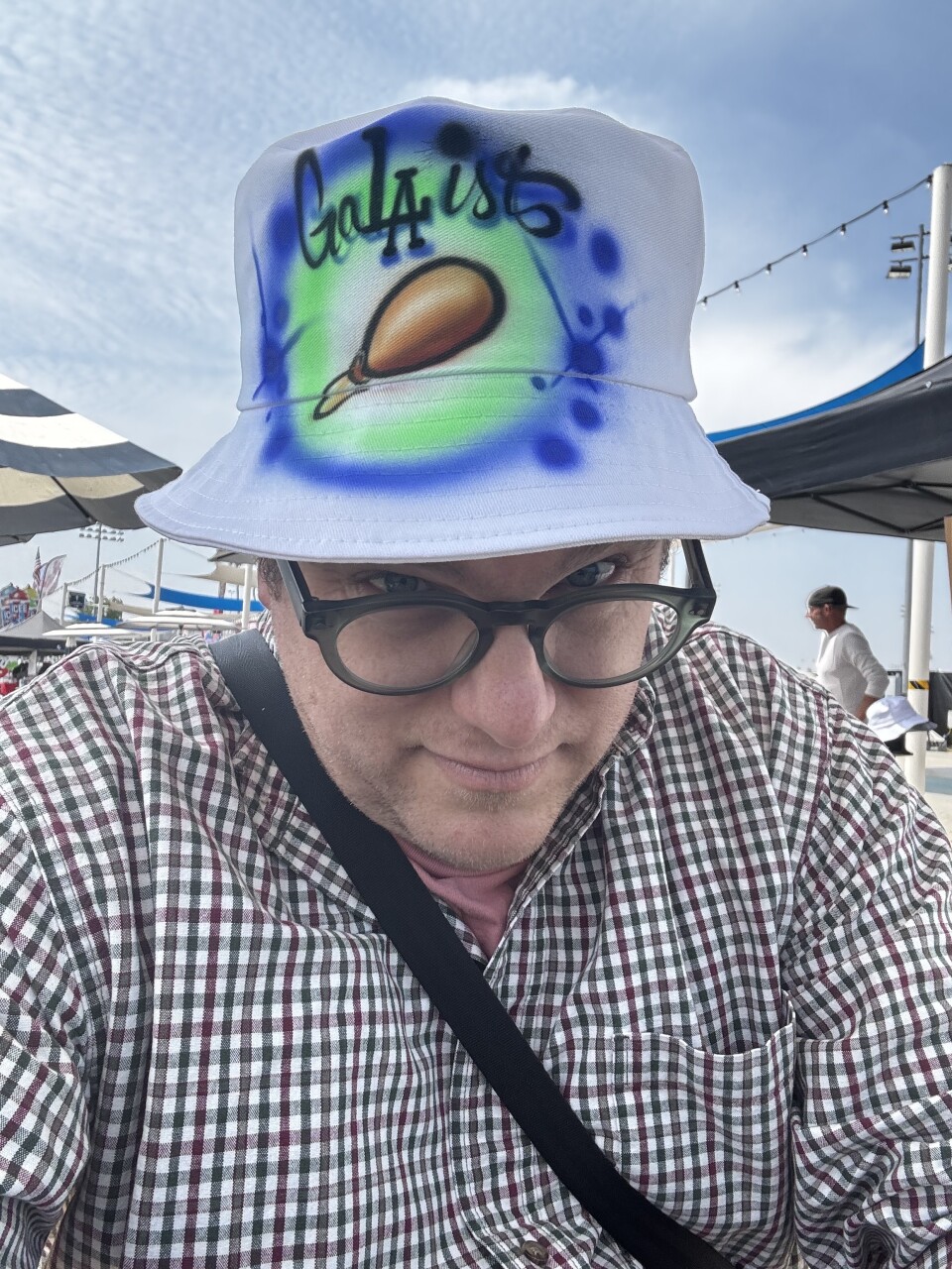 Man with a light skin toned is dressed in a plaid shirt and glasses, is sporting a custom airbrushed bucket hat that reads Ga Laist" in vivid blue and green spray paint design