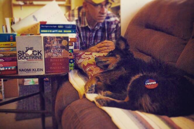 A black and brown dog lays down on a brown sofa on the foreground. In the background, a man wearing a plaid shirt sits. 