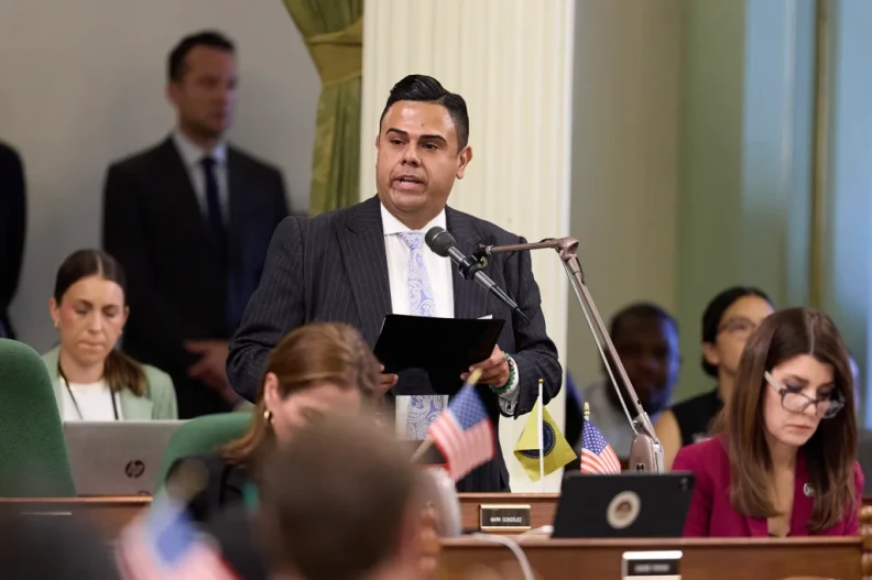 Assemblymember Mark González, a man with medium skin tone, wearing a black stripped suit, speaks into a microphone in a room other people sitting around him.