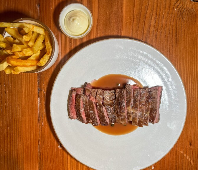 A collection of plates is arranged on a medium dark brown surface. In the upper right corner, there are golden-yellow French fries accompanied by a small container of white sauce that resembles mayonnaise. In the center, a large round white ceramic plate holds slices of cooked brown steak, drizzled with a light brown sauce.