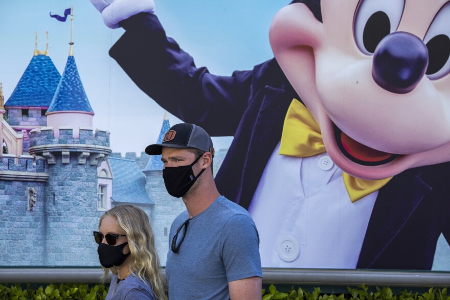 A couple wearing masks walk past a large image of Mickey Mouse as they enter Disneyland.
