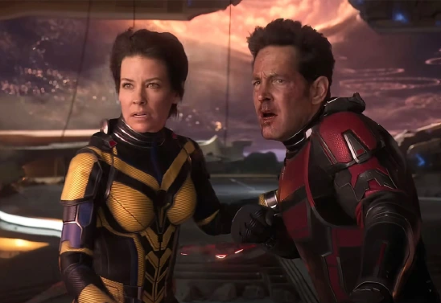 Evangeline Lilly and Paul Rudd stand together in a futuristic space ship. Lilly wears a black and yellow suit that is decoratively segmented into metal armored plates. The suit covers her whole body, only her head visible. Her mouth is slightly opened and brows slightly furrowed looking at someone out of frame. She grasps the right arm of Rudd with her left hand. Rudd wears a similar armored suit but in red and black. His head is also visible. Blood streaks down his face from his nose. He looks flushed with his mouth open and eyebrows pushed together. 