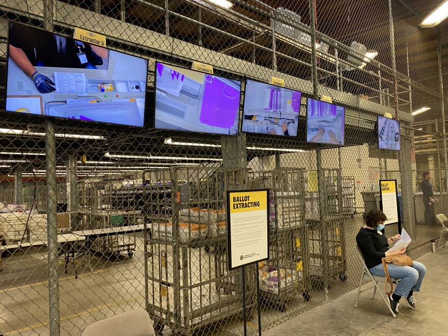 Monitors are attached to a chain-link fence inside a warehouse. The monitors show people handling voting materials.