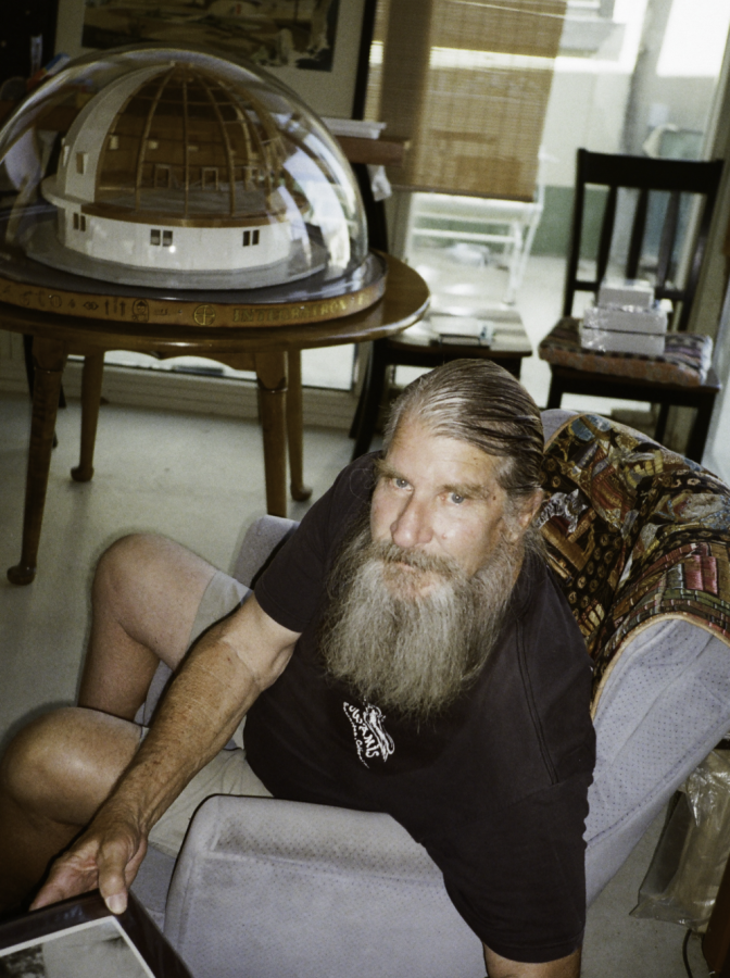 A photo of a long-bearded man sitting in a living room with an architectural model of a domed building sitting on a table behind him. 