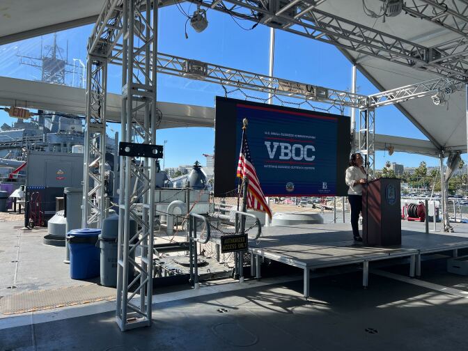 A female presenting person in slacks and a white shirt speaks at a podium in front of a blue screen with the letters VBOC. An American flag is visible on the stage. Scaffolding as well as a battle ship is visible in the background. 