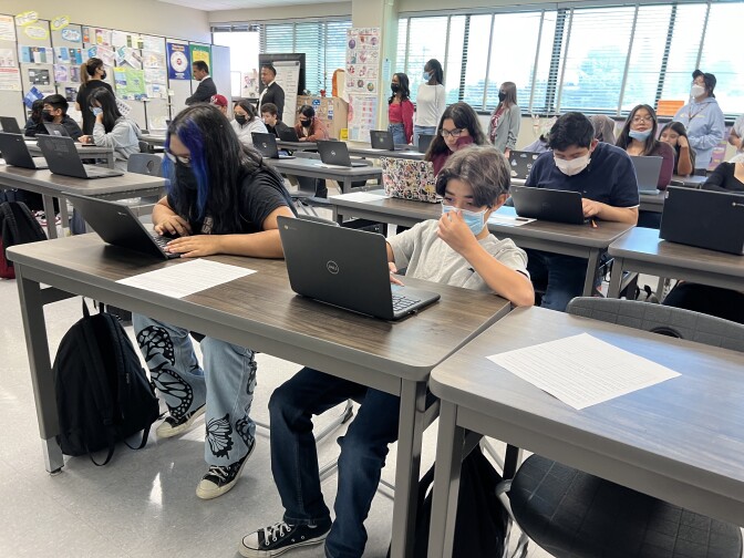 A classroom full of teenagers sit at long tables and stare at laptop screens in a classroom.