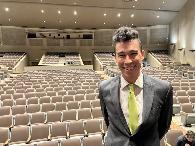 A man in a suit smiling on stage with concert seating behind him.