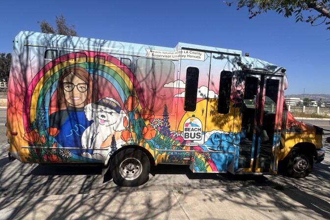 A colorful half-size passenger bus is parked next to a sidewalk. It has the image of a sunset, a blue ocean, and a woman wearing glasses carrying a white dog with a hat that says "WINSTON".