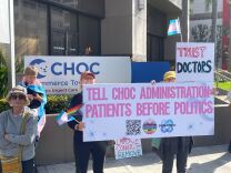 Protesters holding trans and LGBTQ+ pride flags pose for a photo outside Children Hospital of Orange County. Their signs say "Tell CHOC administration: Patients before politics," "Impeach, convict, and remove," and "Trust doctors."