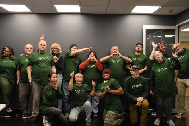 A group of more than a dozen people wearing green 'fountain house' t-shirts pose for a photo. Many are smiling and some make heart and thumbs up gestures with their hands. 