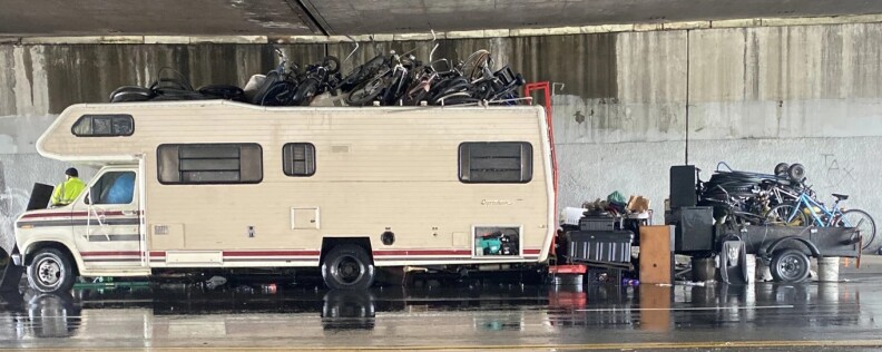 A white RV with red trim is parked under a freeway overpass with bicycles piled on top of it and on a trailer behind it.